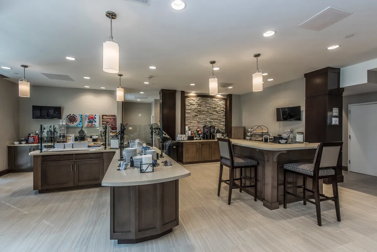 A bright interior coffee bar and snack station with a modern seating area. The setup includes coffee dispensers, various breakfast items, and inviting chairs for guests to relax and enjoy refreshments or socialize.