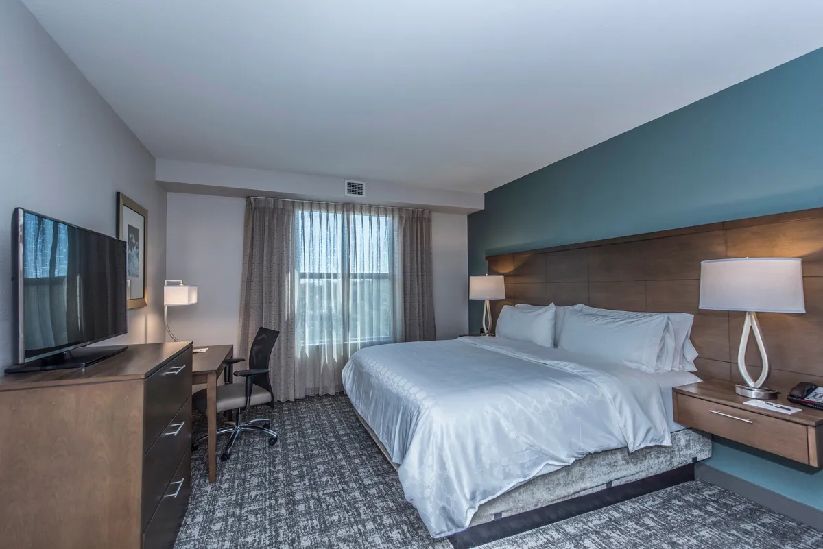 A spacious king-sized bedroom featuring a large window draped with sheer curtains, a plush bed neatly made with fresh linens, and a dedicated desk area with a chair. A television rests atop a dresser, while tasteful art and soft lighting create a tranquil atmosphere ideal for rest and productivity.