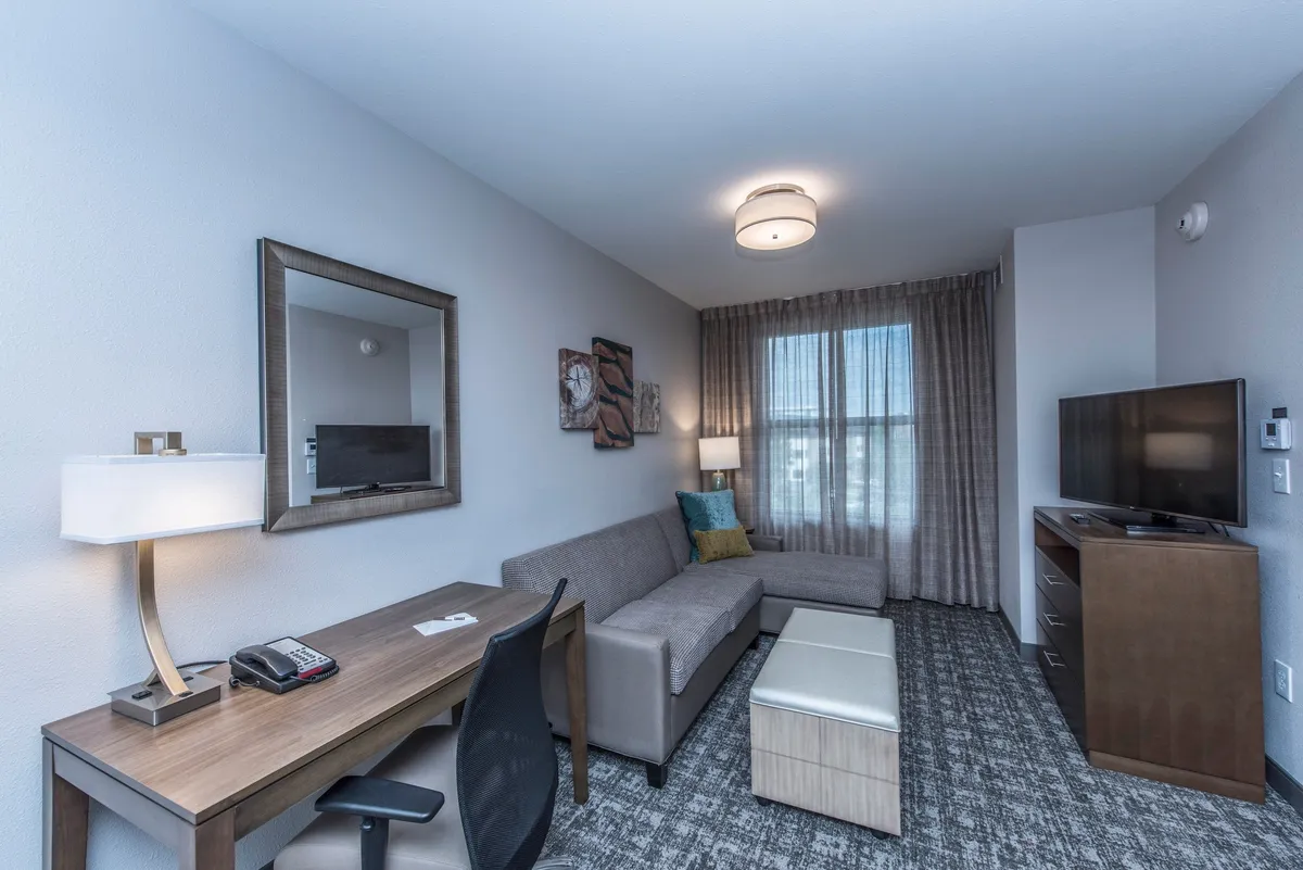 A contemporary living area with a comfortable sofa, a work desk with a chair, a television mounted opposite the seating area, and decorative art adorning the walls. Soft lighting from a lamp and touches of modern decor create a welcoming space for relaxing or working.