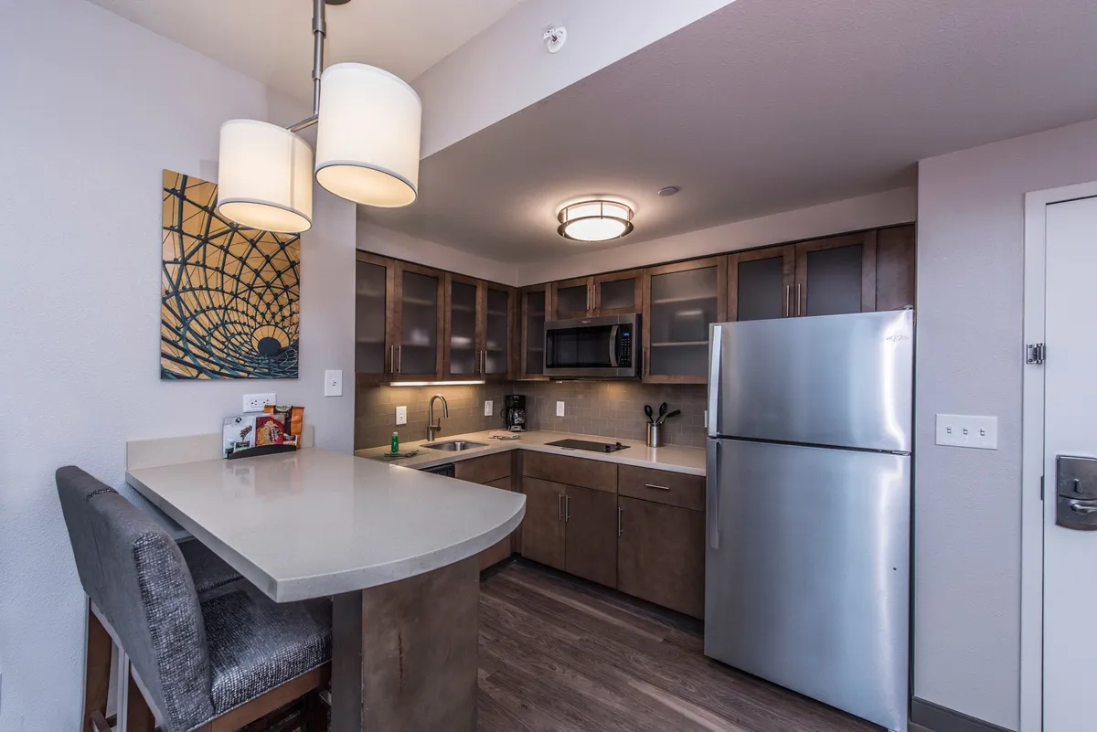 A spacious, modern kitchen featuring a large island, stainless steel refrigerator, contemporary lighting fixtures, and decorative wall art. The space is designed for both cooking and entertaining, with functional counter space and seating that invites guests to share a meal or gather for casual dining.