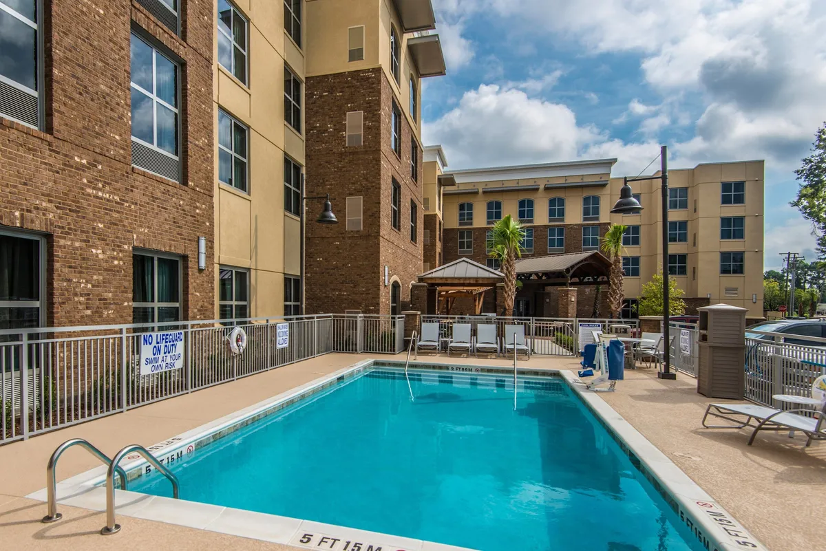This image features a sparkling outdoor swimming pool surrounded by palm trees and an array of comfortable lounge chairs. The building's architecture frames the scene, and the area exudes an inviting, resort-like feel ideal for relaxing or socializing under the sun or stars.