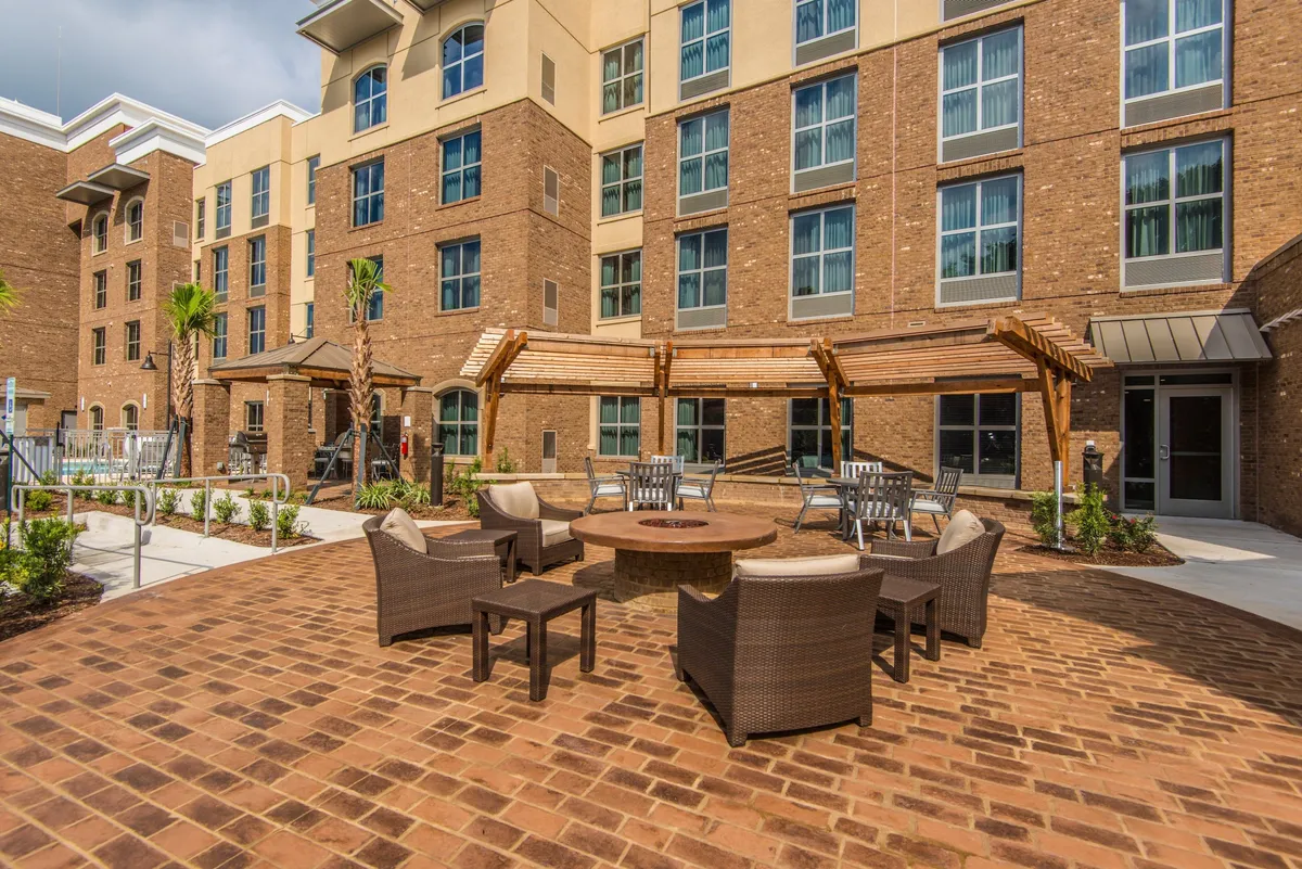 An inviting exterior scene featuring a cozy fire pit surrounded by comfortable seating under palm trees, with the hotel facade as a backdrop. The space appears ready for social gatherings, relaxation, or informal networking during the evening, encapsulating the retreat-worthy amenities available to guests.