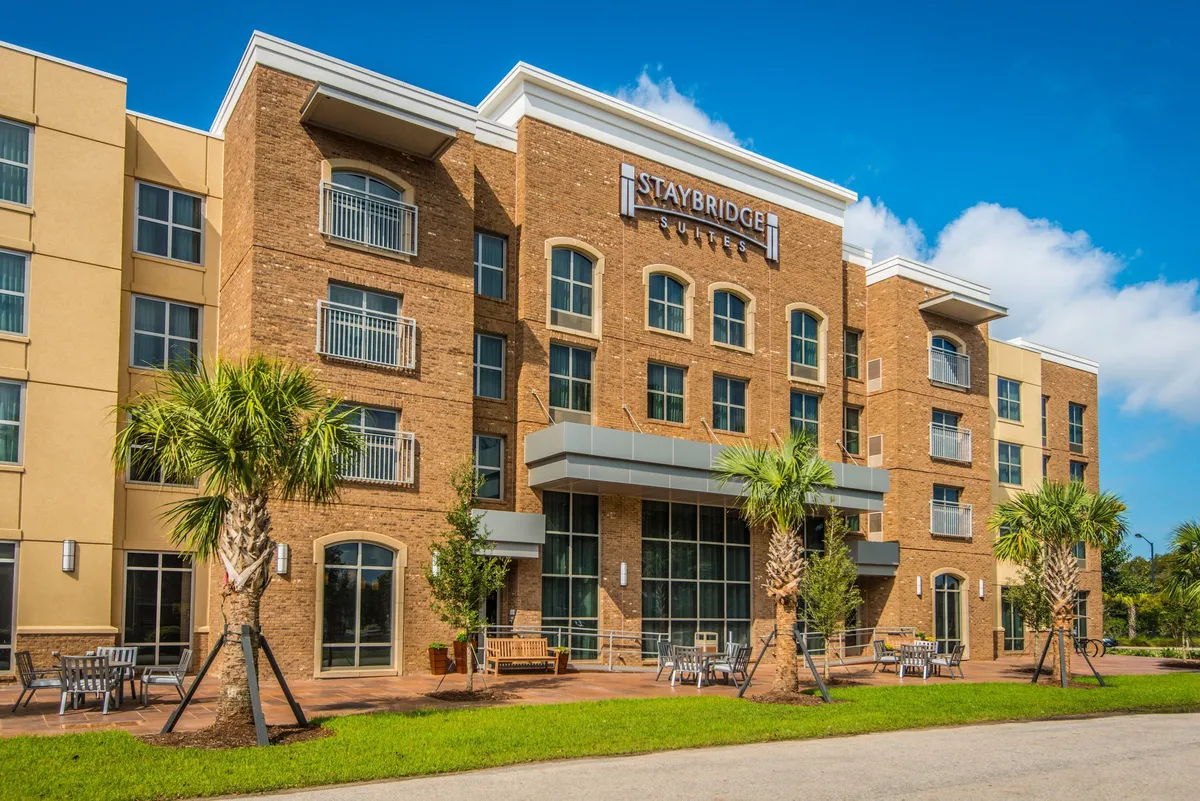 This exterior view showcases the Staybridge Suites signage, towering palm trees, and a welcoming outdoor seating area bathed in natural light. The setting encourages guests to enjoy coffee in the morning or gather for sunset views.