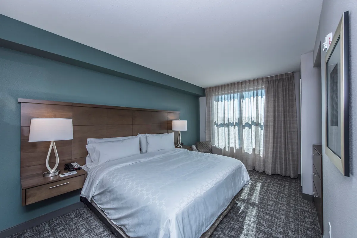 A tranquil, comfortable suite interior featuring a plush bed with lamps on either side, soft curtains filtering sunlight, and an inviting armchair in the corner—perfect for unwinding between festival events.