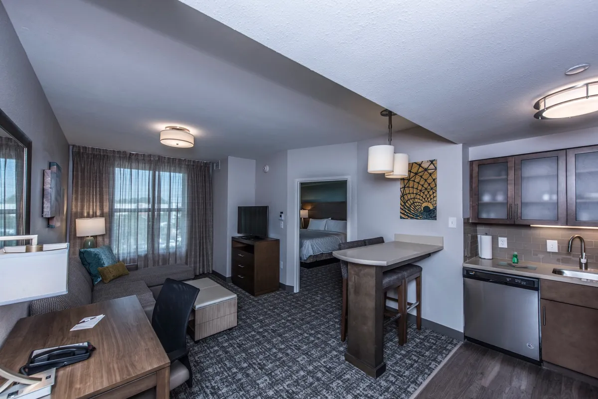 The image showcases an inviting condo interior with a comfortable seating area, a modern kitchenette, a functional work desk, and a glimpse into the bedroom. The setting is warm and welcoming, perfect for relaxing, working, or sharing meals.